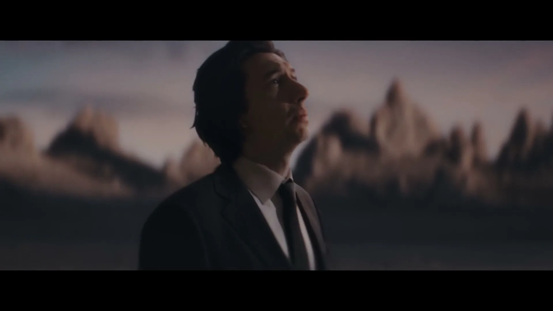 Adam Driver, a male in his 30s, wearing a dark suit, looks up and then down, touching his forehead in a vast, desolate landscape.