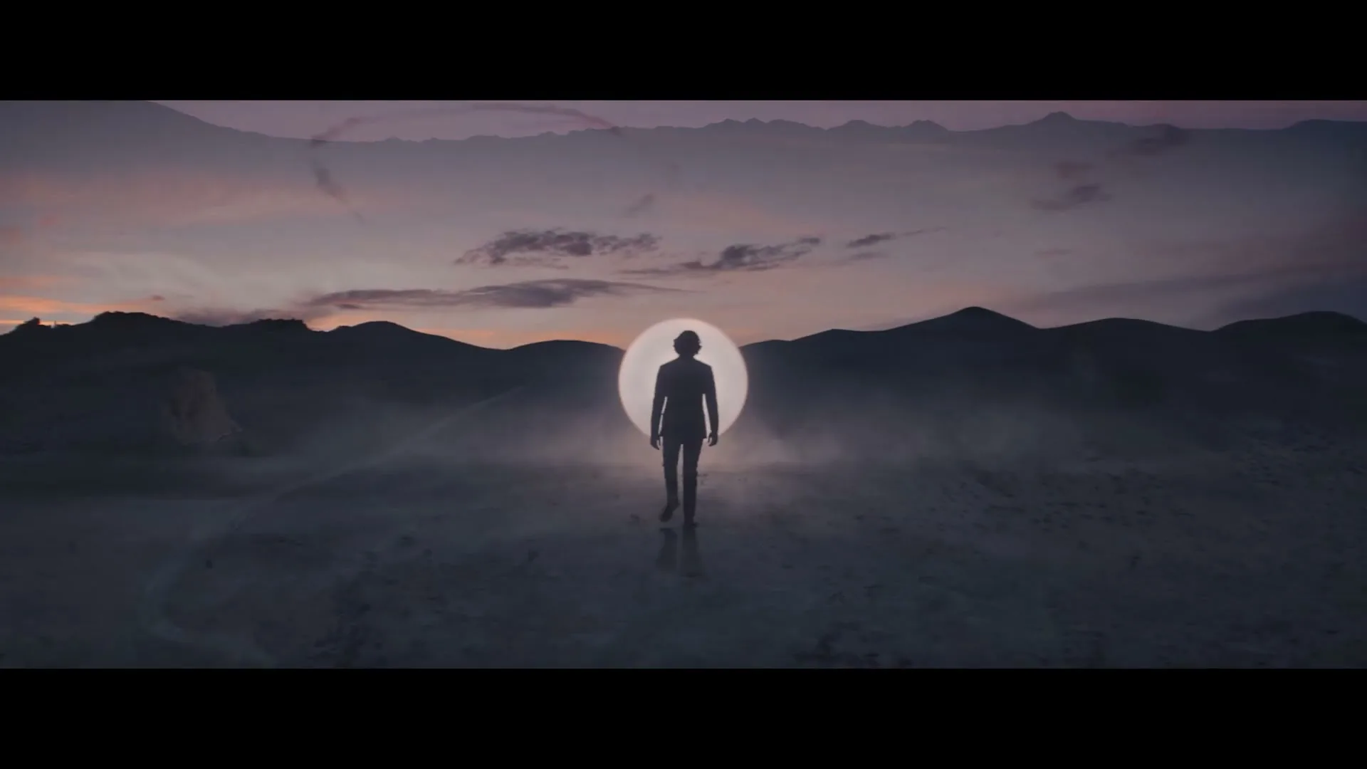 A wide shot of a person standing in a vast desert landscape with rock formations, silhouetted against a glowing circle and a colorful sky at dusk.