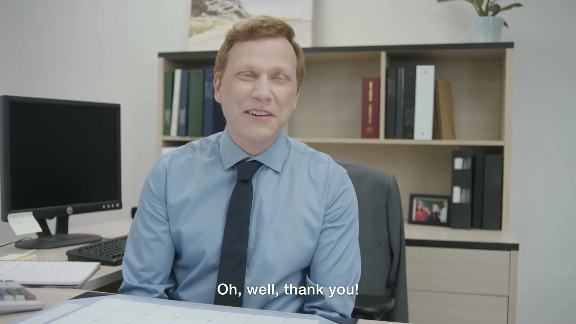 A man with red hair and a blue shirt sits at a desk in an office, looking slightly uncomfortable as he responds to someone off-screen.