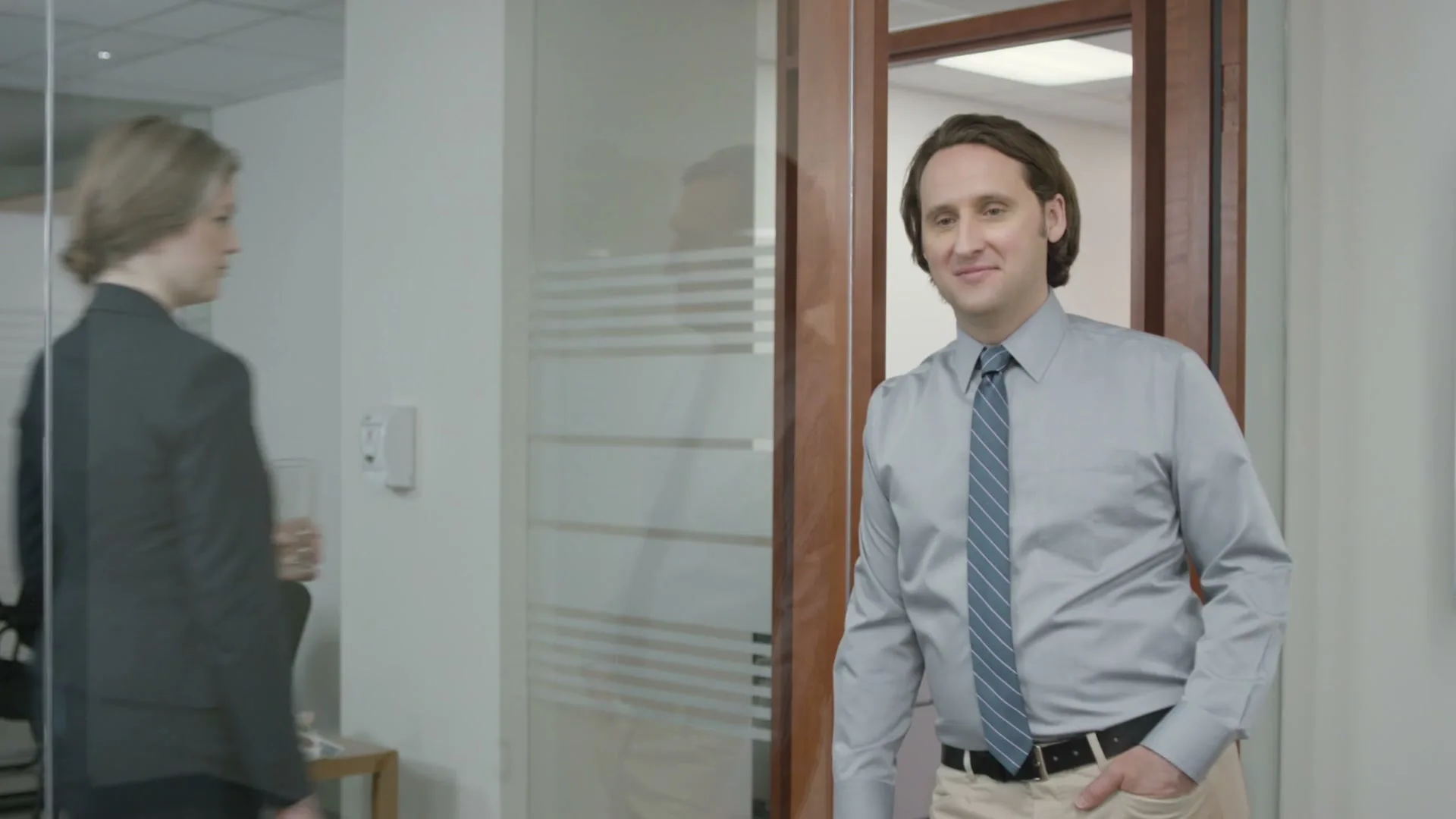 A man in a shirt and tie stands in an office doorway, smiling, as a woman walks past him in the background.