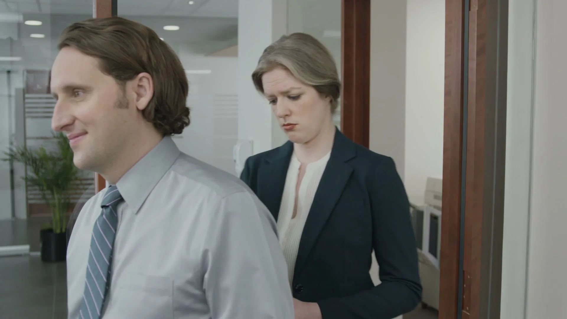 A man in a dress shirt and tie smiles, while a woman in a business suit behind him looks displeased in an office setting.