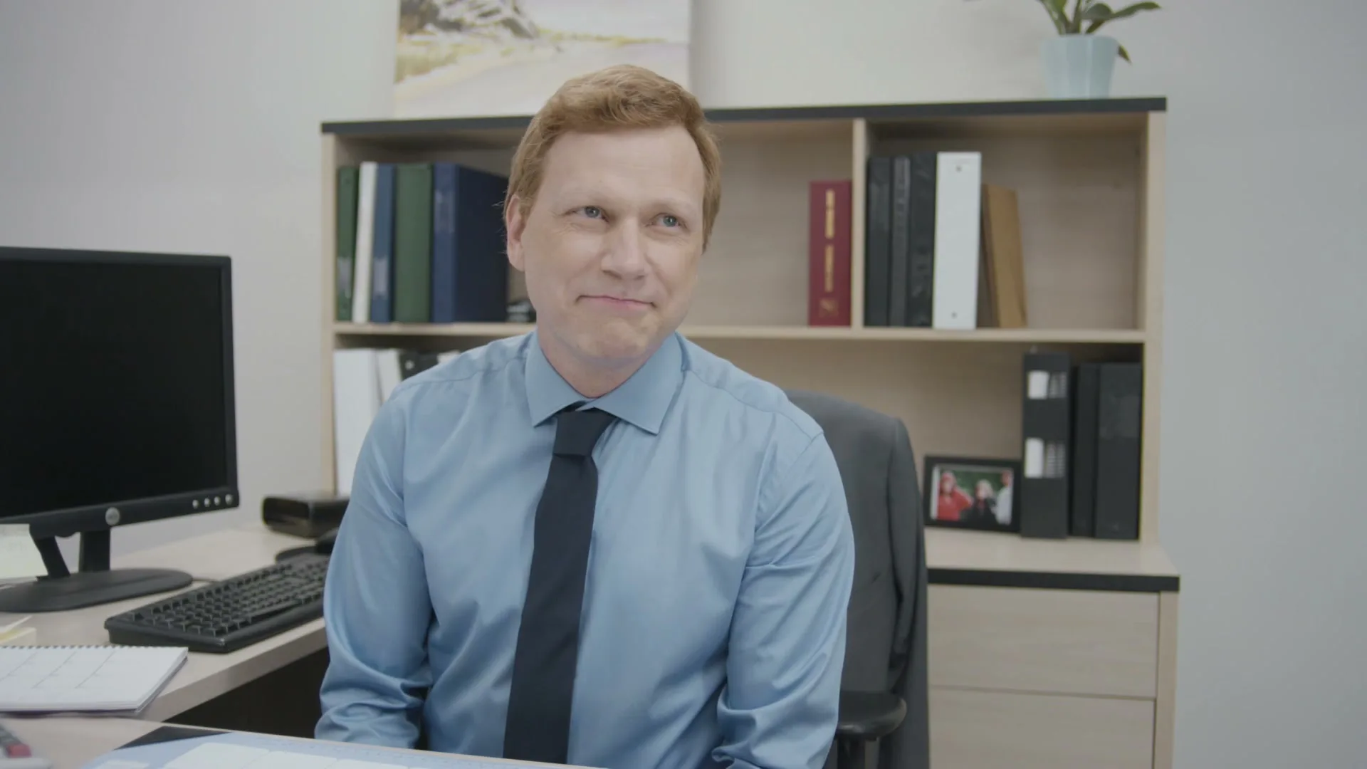 A man with red hair in a blue shirt and tie sits at an office desk, looking slightly to the right with a subtle smile.