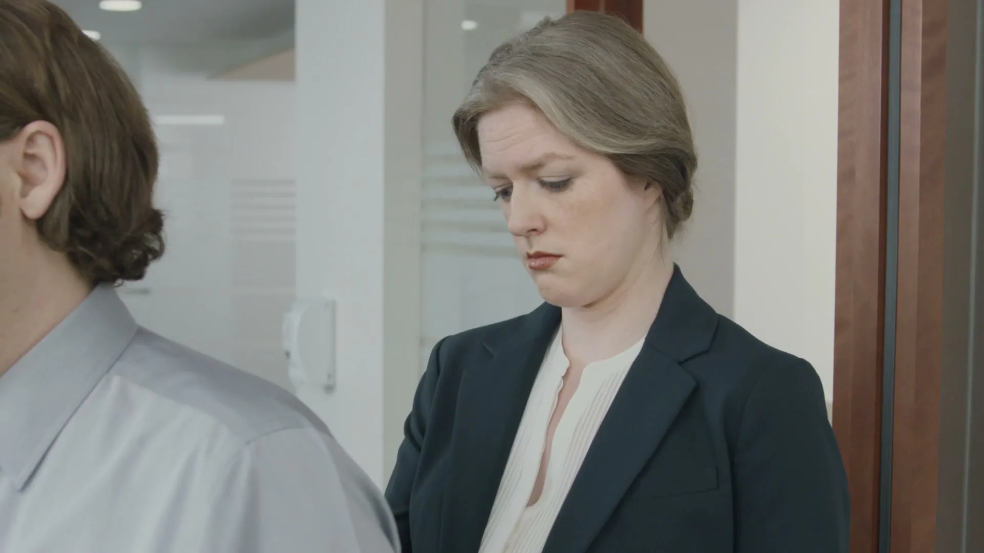 A woman with grey hair in a dark suit looks down with a concerned expression, standing next to a man in a grey shirt in an office.
