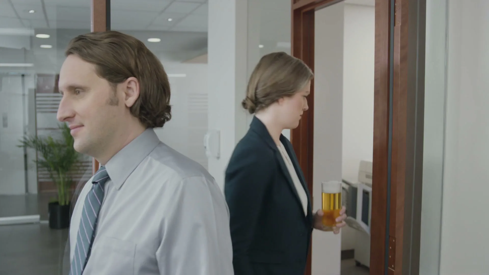 A man in a dress shirt and tie smiles as a woman in a business suit walks past him carrying a glass of beer in an office hallway.