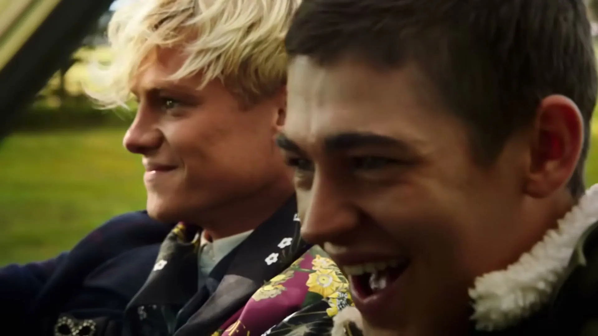 Two young men, one with blonde hair and the other with brown hair, are riding in a car, smiling and laughing joyfully.