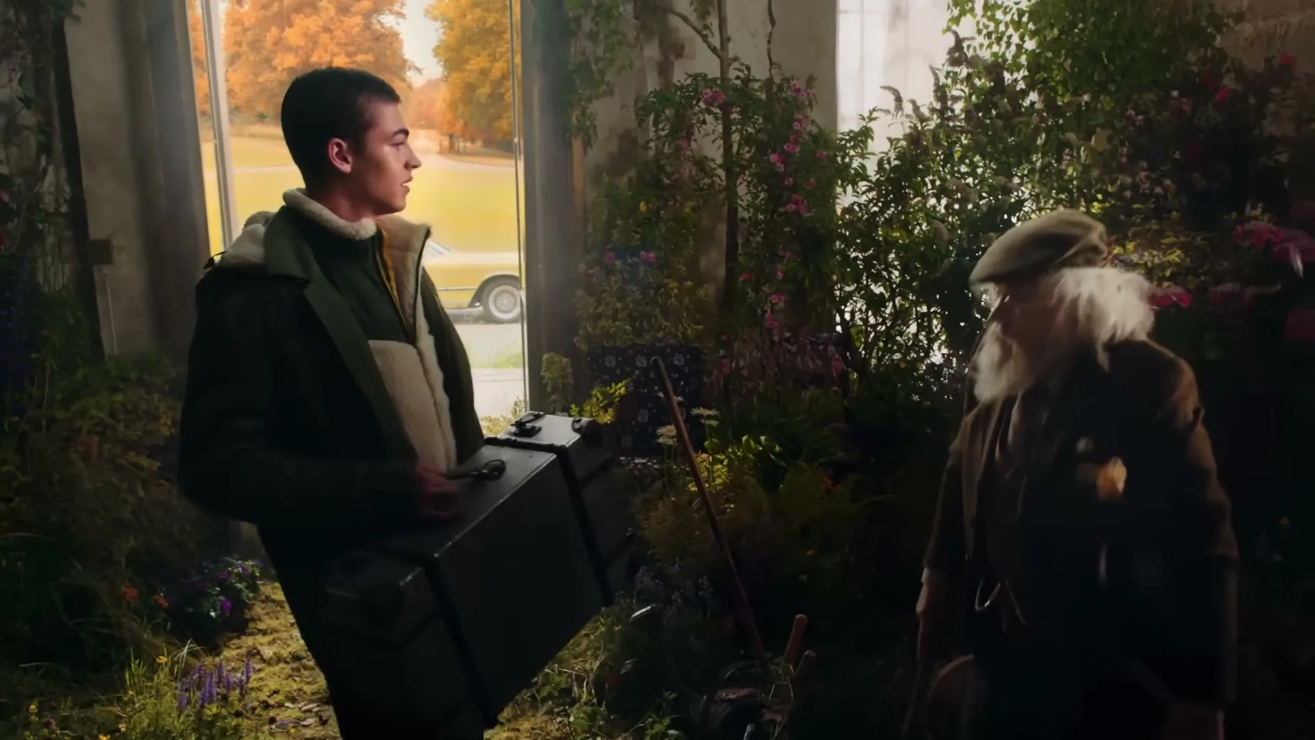 A young man in a green jacket and an older man with a white beard stand in a room overgrown with plants, looking at each other.