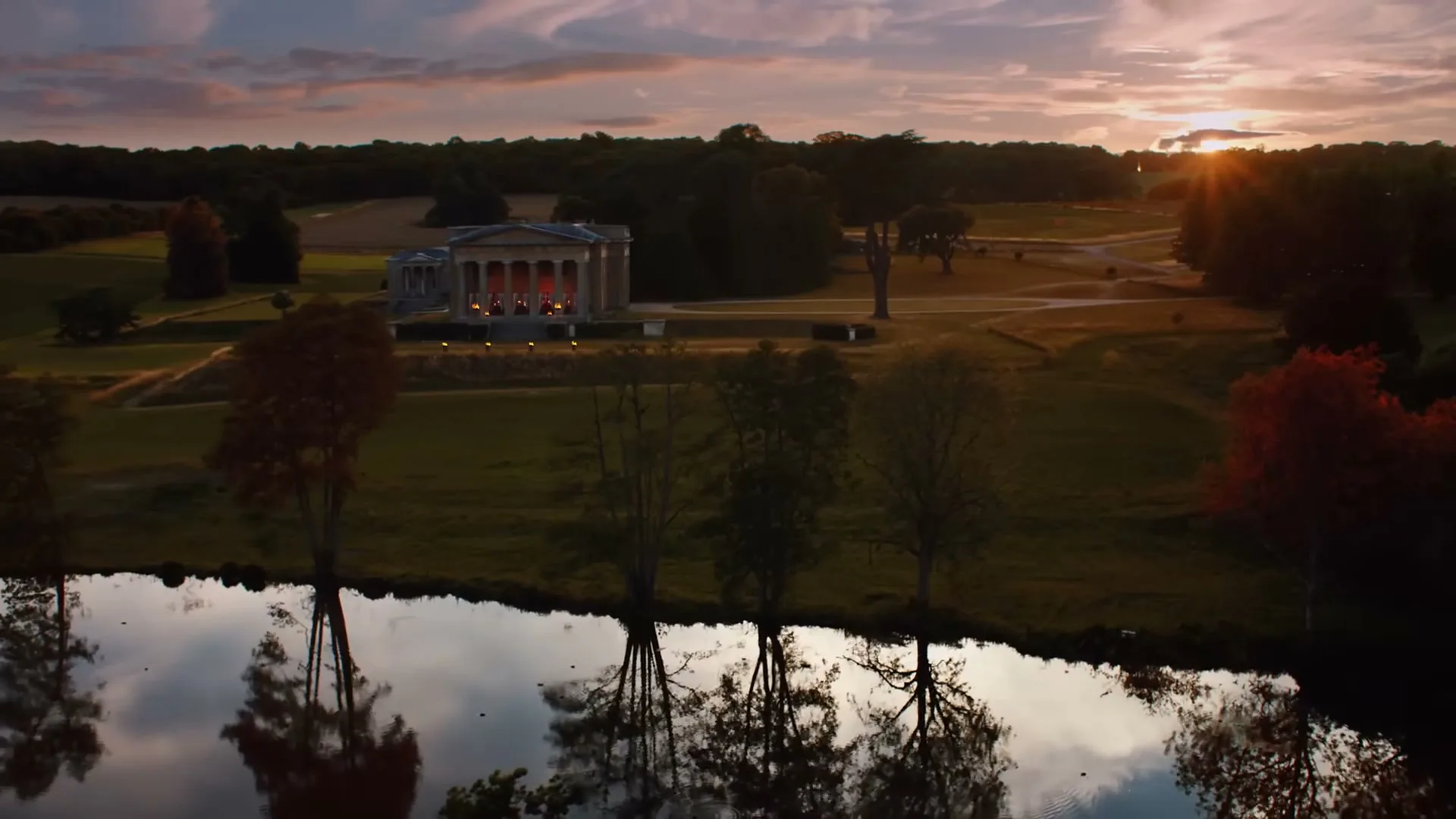 An aerial view of a grand classical building with illuminated columns, surrounded by expansive green fields and trees, with a reflective body of water in the foreground at sunset.
