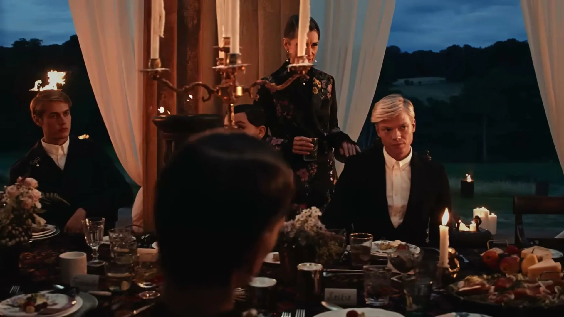 A woman in a floral jacket stands behind a table, looking at a young man with blonde hair, at an outdoor dinner party.
