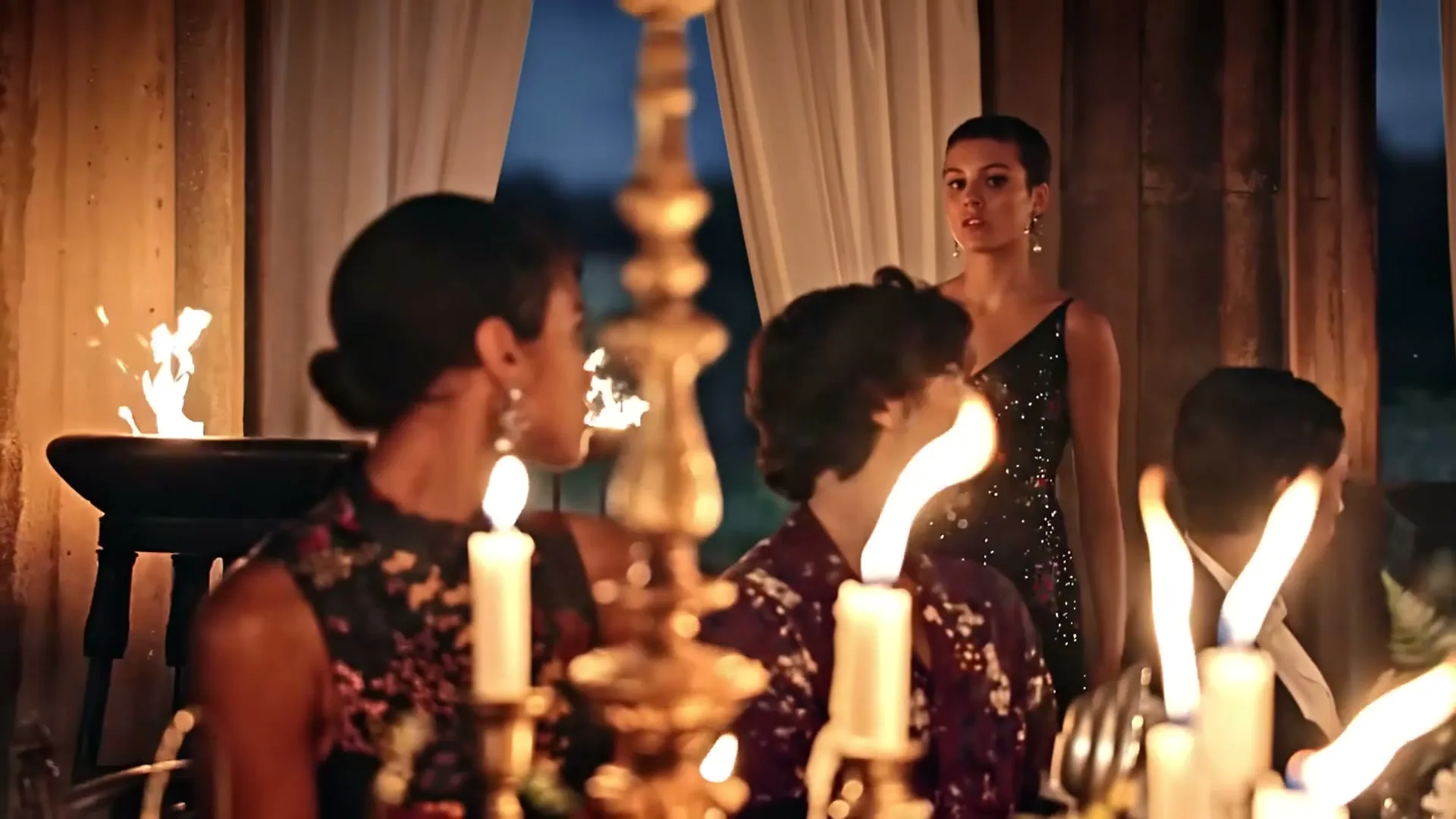 A young woman with short hair and a black dress stands at a candlelit dinner table, looking intently at someone off-screen, surrounded by other guests.
