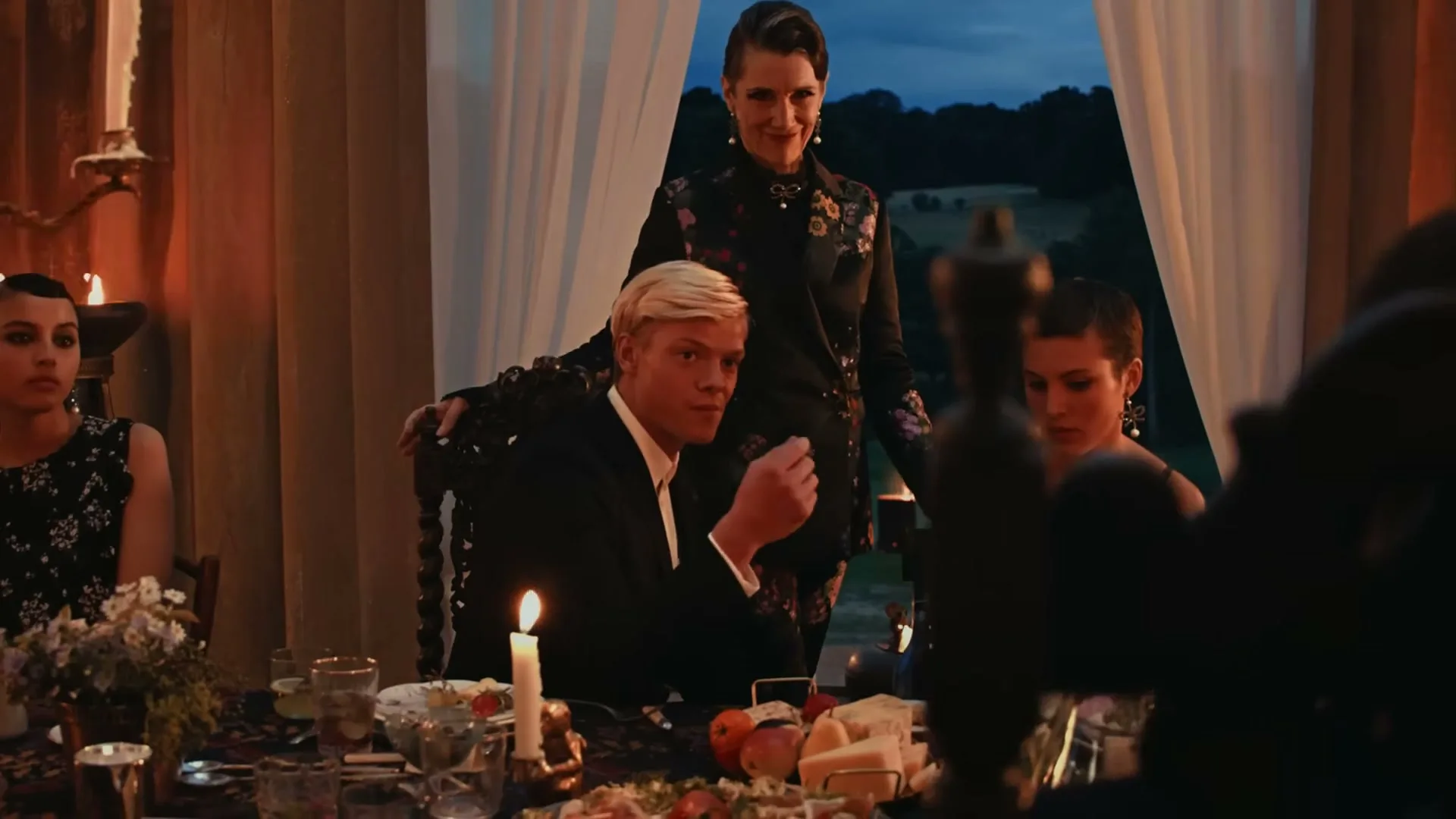 A woman in a floral jacket stands behind a man with blonde hair at a candlelit dinner table, while another woman sits to the right, in a grand dining room.