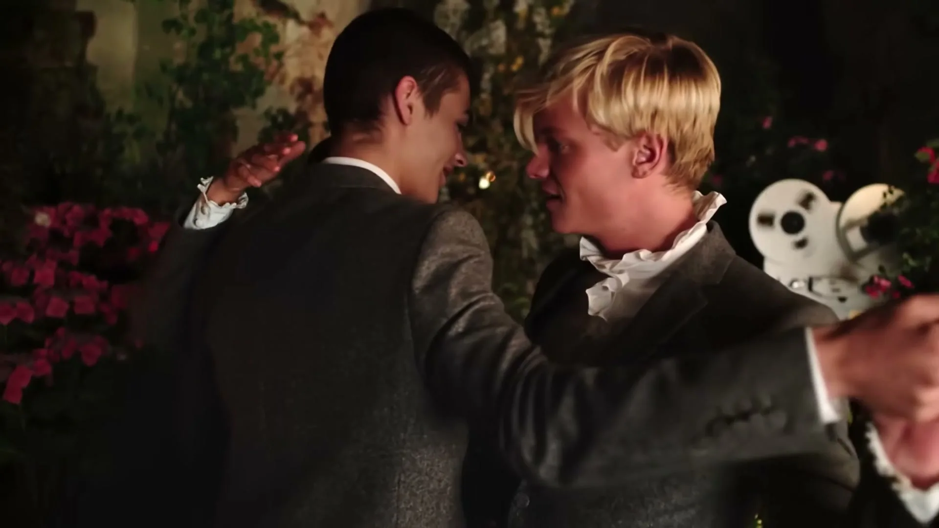 Two young men in period suits dance closely, one with blonde hair smiling at the other, in a dimly lit garden party setting.