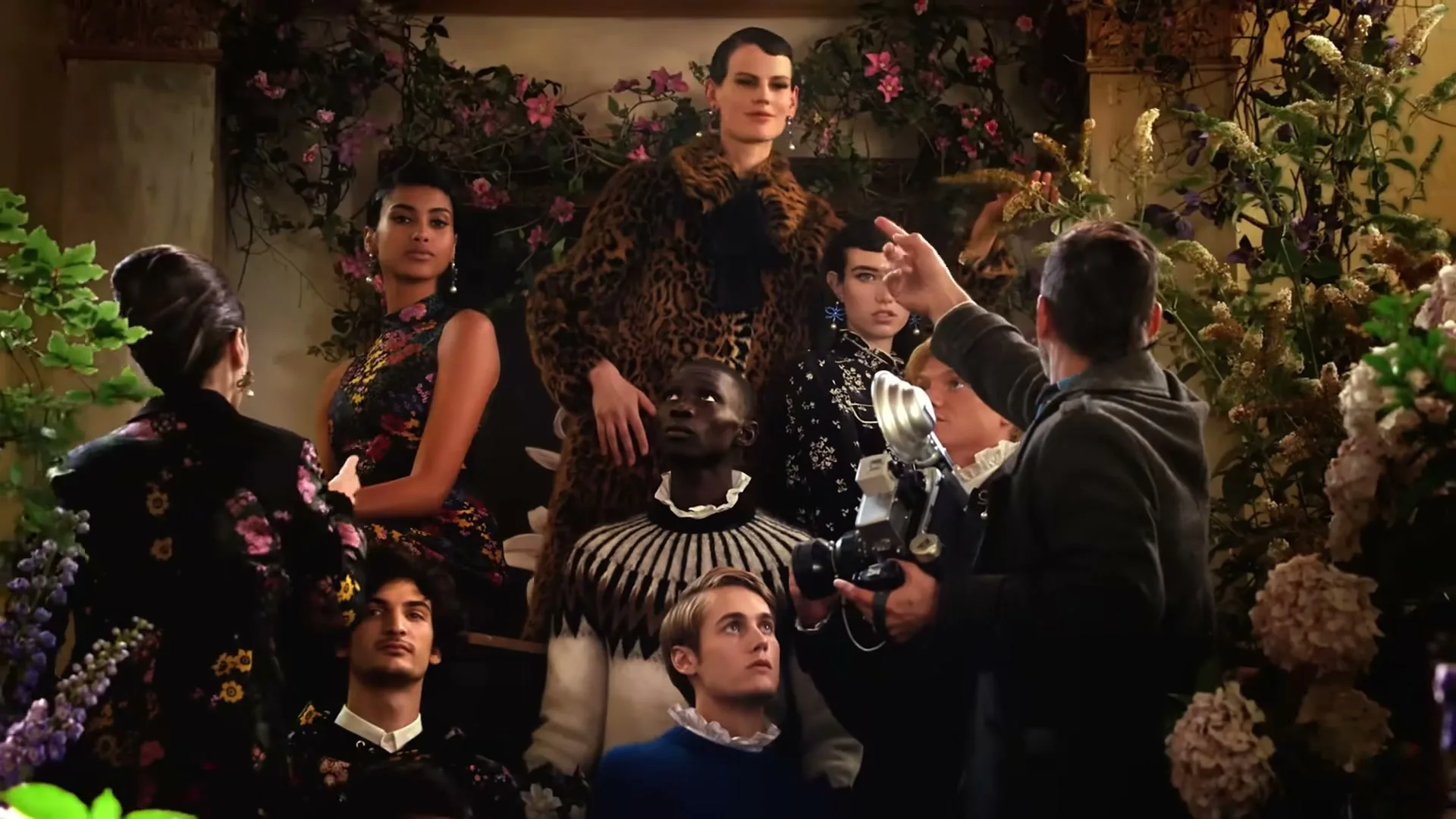 A group of diverse models in elaborate floral outfits pose for a photographer in a lush, flower-filled interior setting.