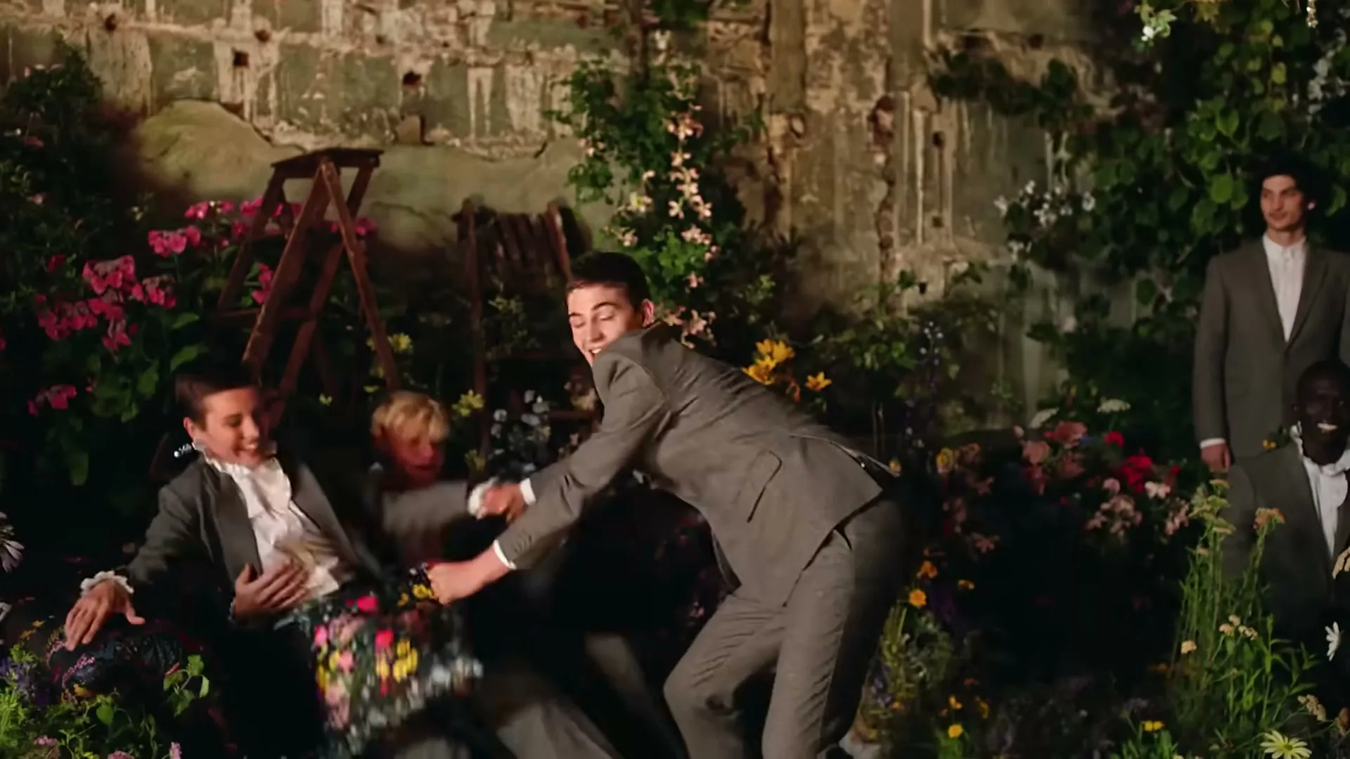 Three young adults in grey suits and white ruffled shirts playfully wrestle over a floral pillow in a lush, overgrown garden setting with stone walls.