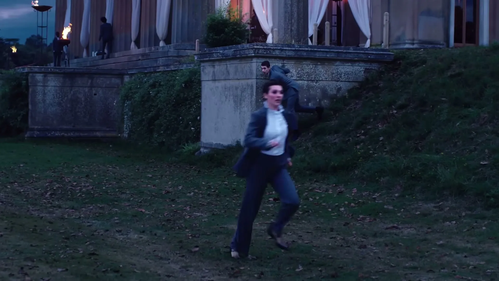A woman in a suit runs across a grassy lawn away from a large building, while a man in a suit peeks from behind a wall.