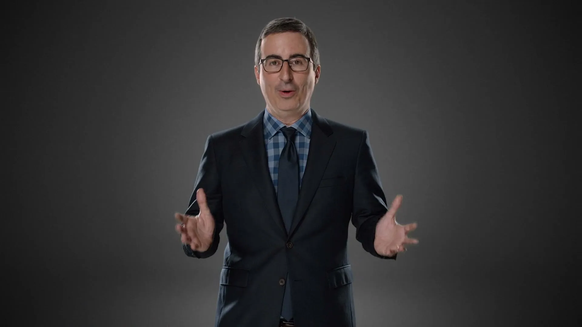 John Oliver, a white man in his 40s, wearing a dark suit and glasses, stands against a gray background, gesturing with his hands as he speaks.