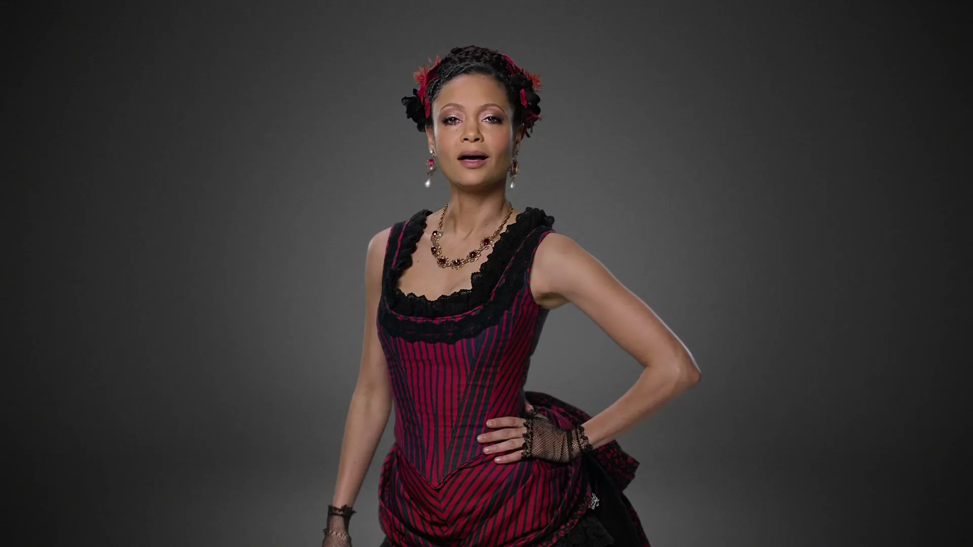 A woman in a red and black striped dress with lace details and a matching headpiece stands against a gray background, looking directly at the camera.
