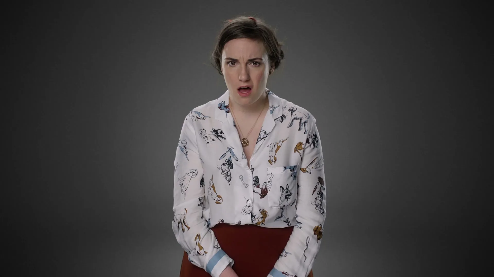 A young woman with dark hair, wearing a white shirt with dog patterns and a red skirt, looks surprised in a studio.