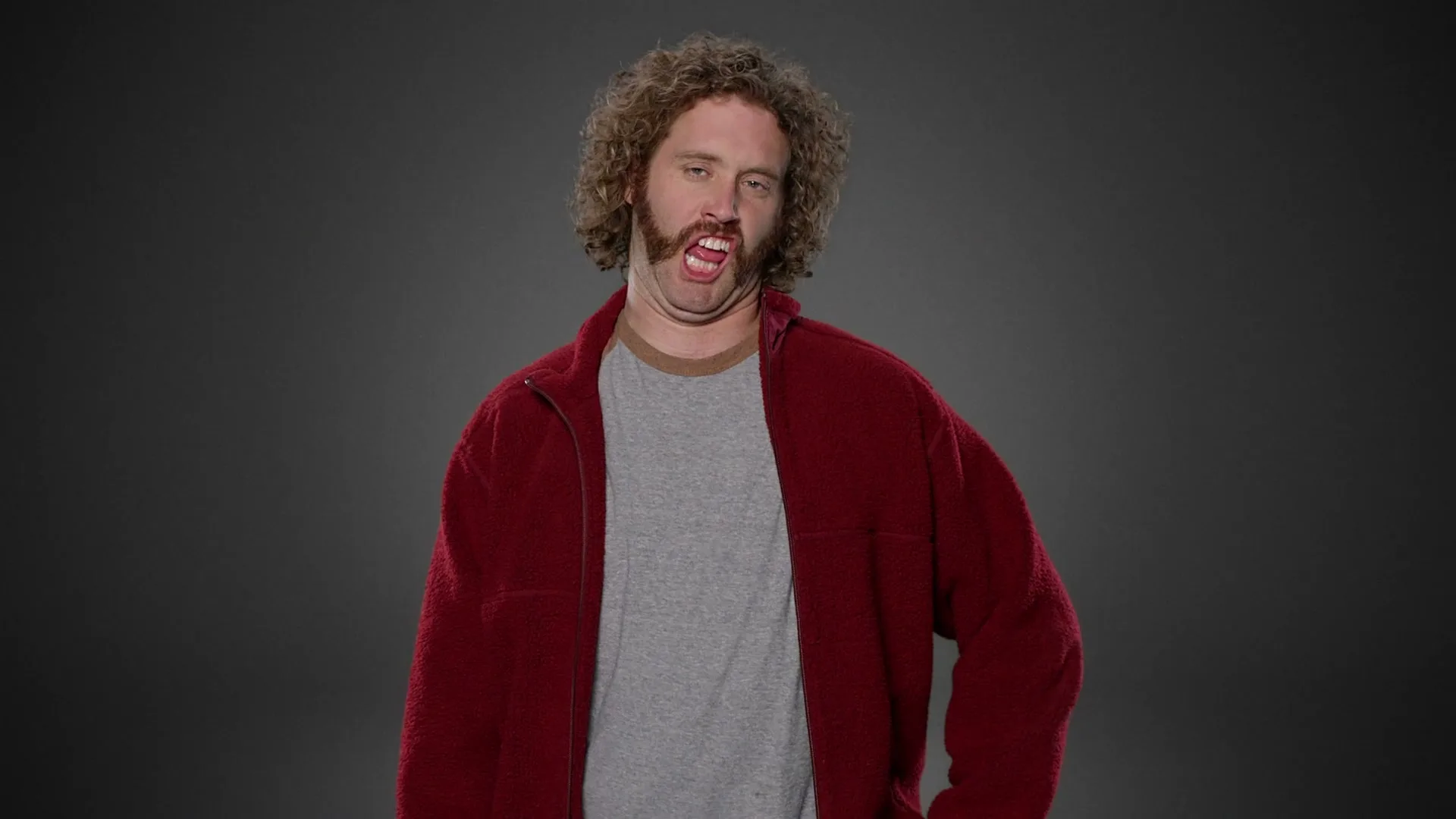 A man with curly hair and a mustache wearing a red jacket and grey t-shirt makes a funny face in a studio.