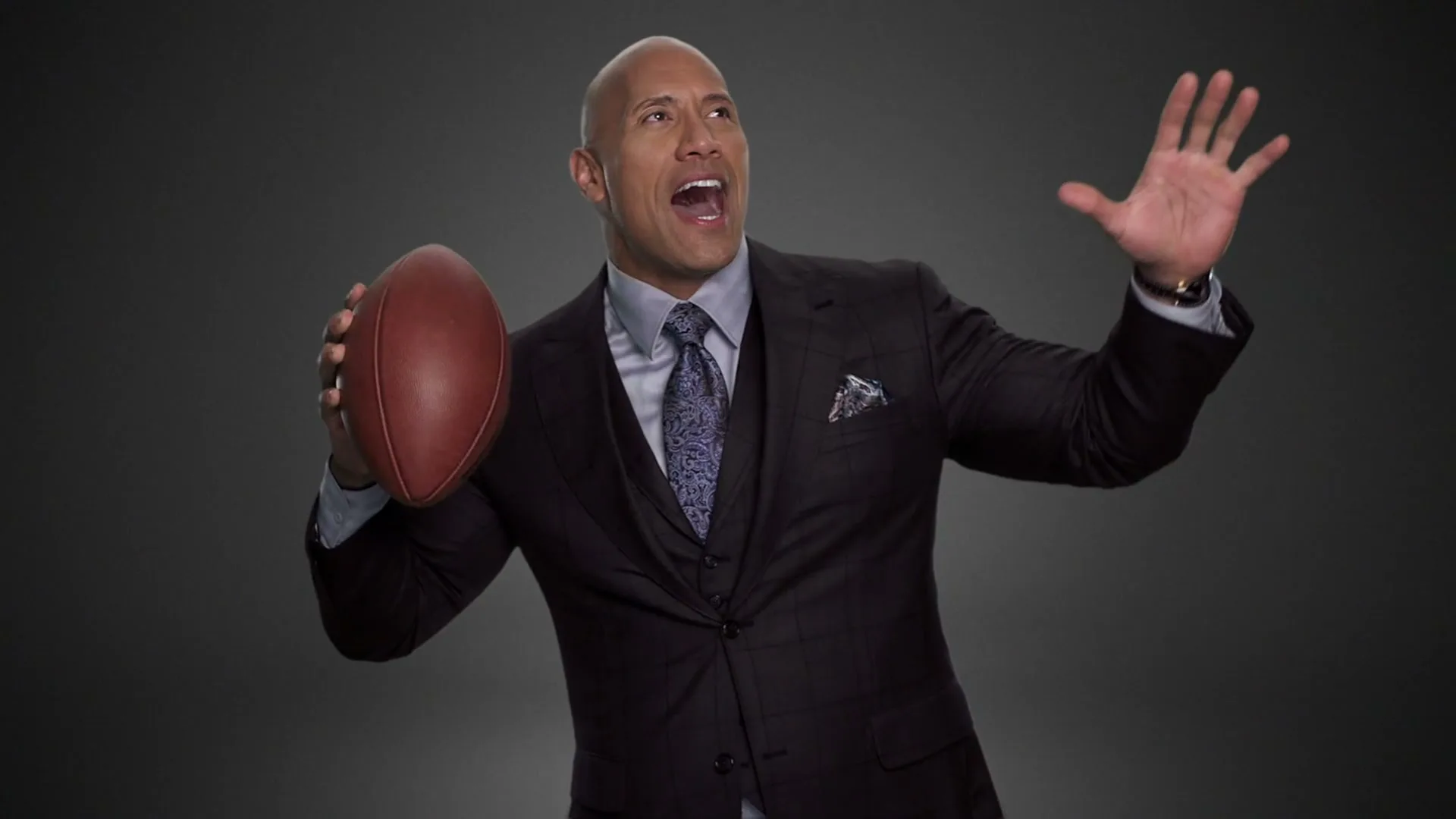 Dwayne "The Rock" Johnson, a bald man in a suit, holds a football and gestures excitedly in a studio.