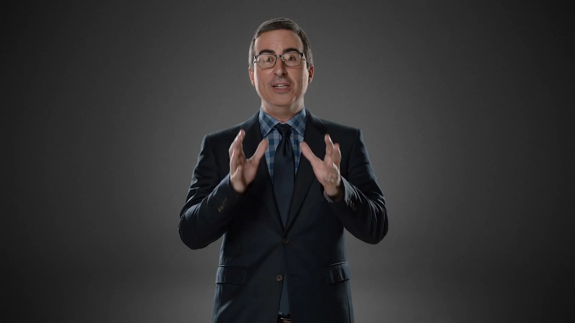 John Oliver, a male in his 40s, wearing a suit and glasses, stands against a gray background, gesturing with his hands as he speaks.