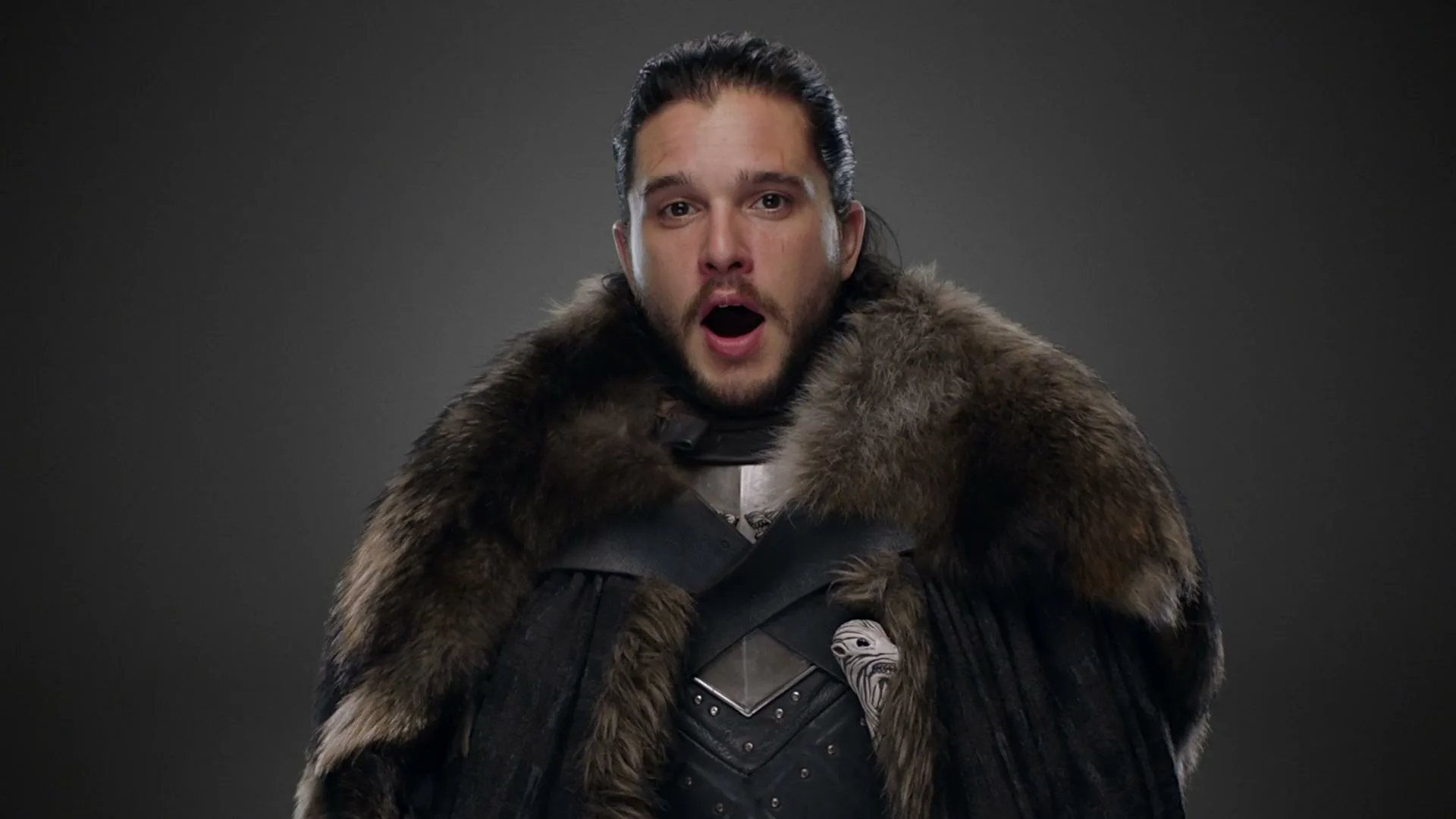 A man with dark hair and a beard, dressed in a fur-lined cape and armor, looks surprised with his mouth open in a studio.