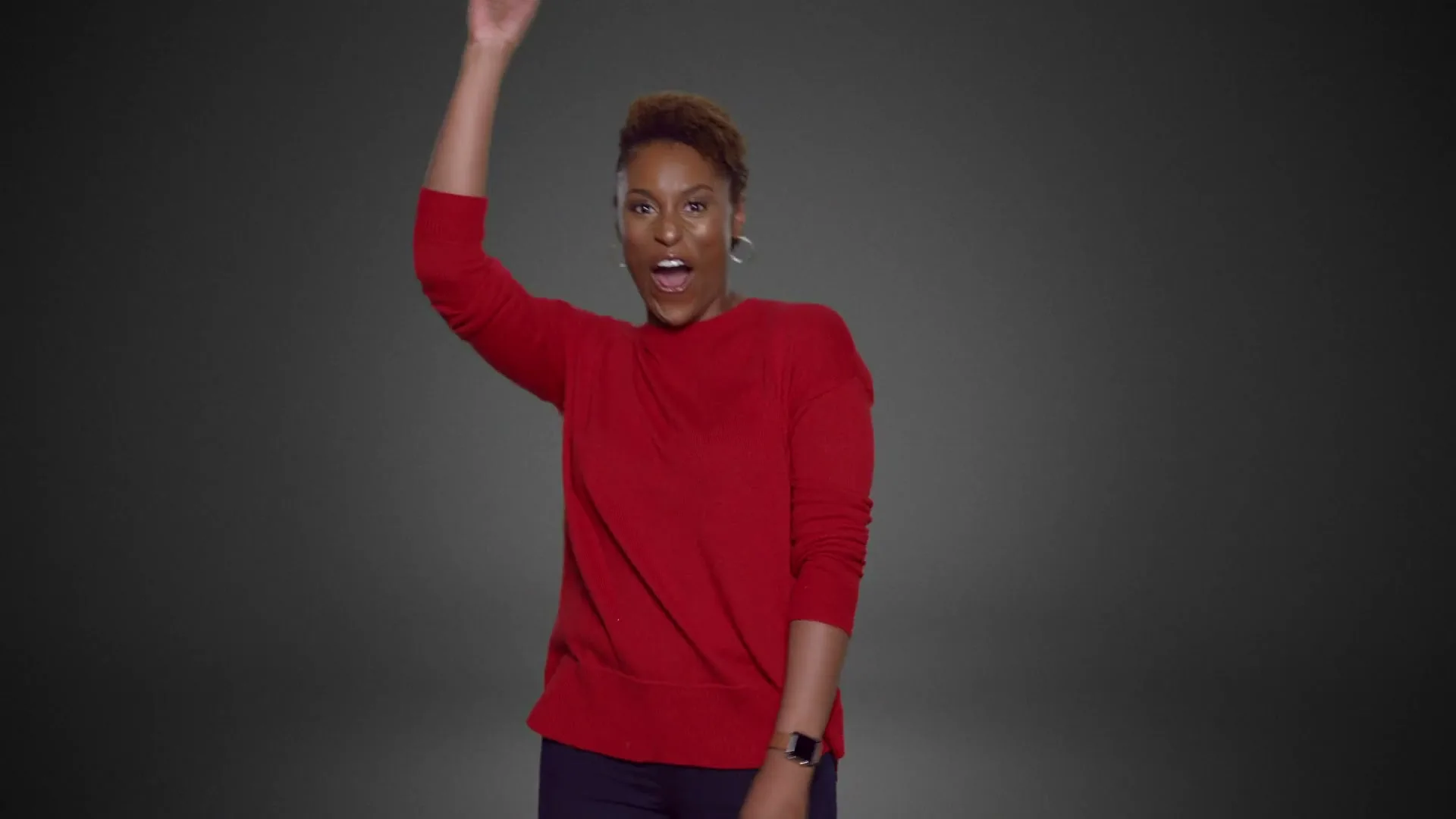 A woman with short brown hair, wearing a red sweater and dark pants, raises her arm and shouts excitedly in a studio.