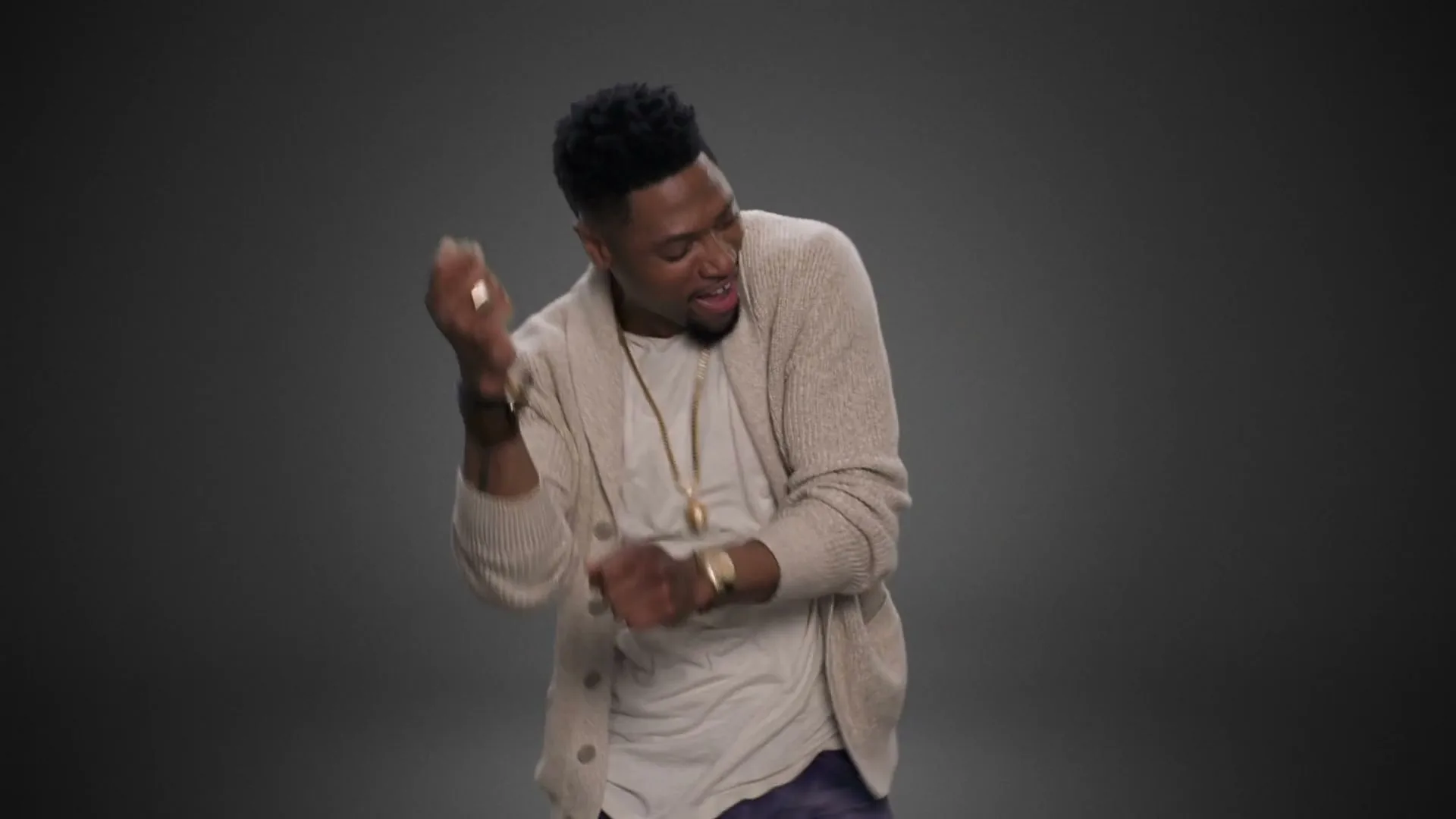 A man with dark skin and hair, wearing a beige cardigan, dances energetically with a joyful expression in a studio.