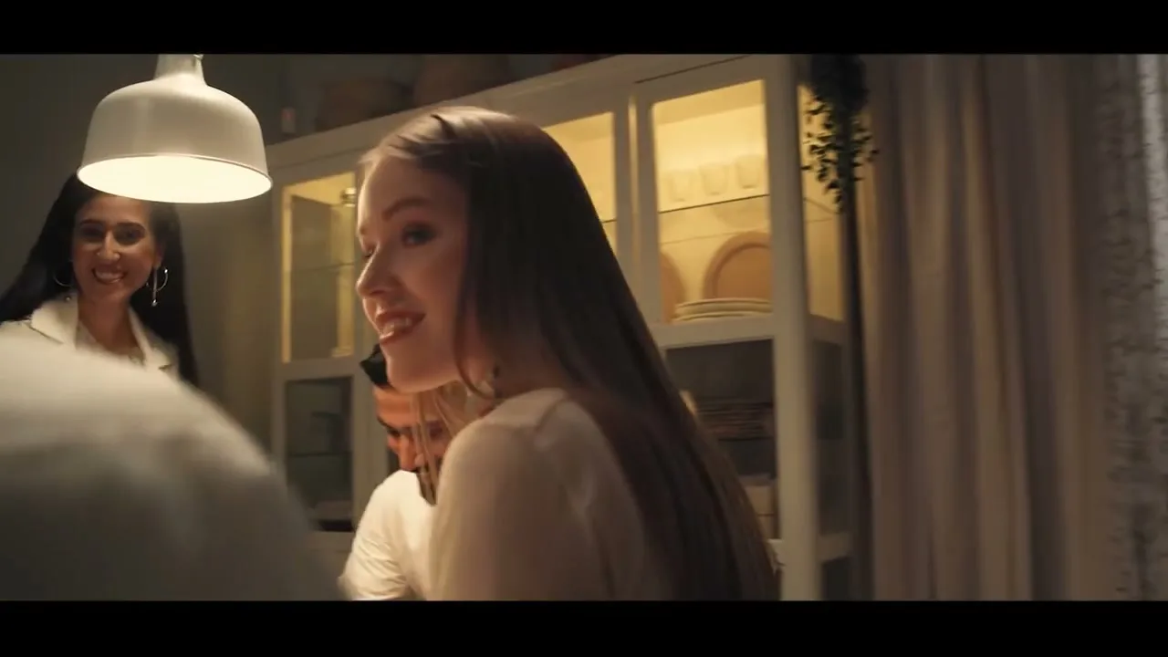 A young woman with long brown hair smiles and looks over her shoulder at a gathering in a dining room, with another woman laughing in the background.
