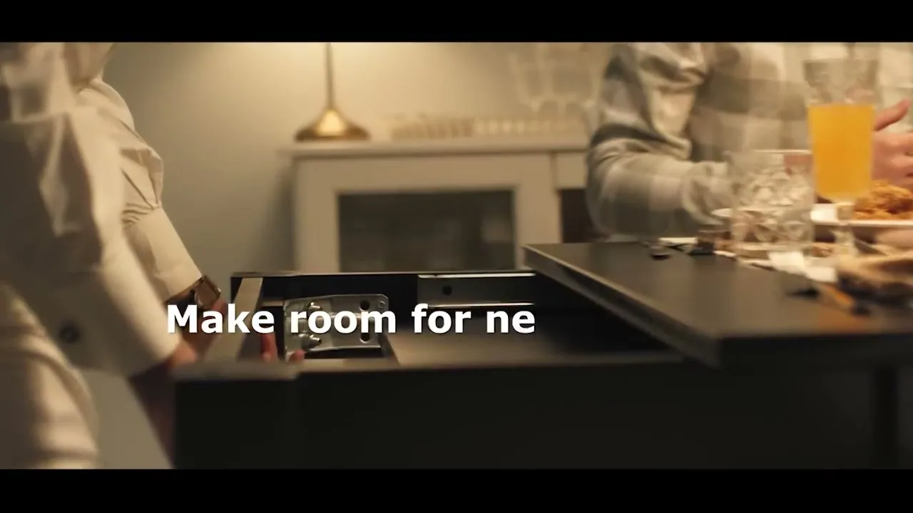 A person in a white shirt extends a black dining table, making room for more friends, in a dimly lit interior.