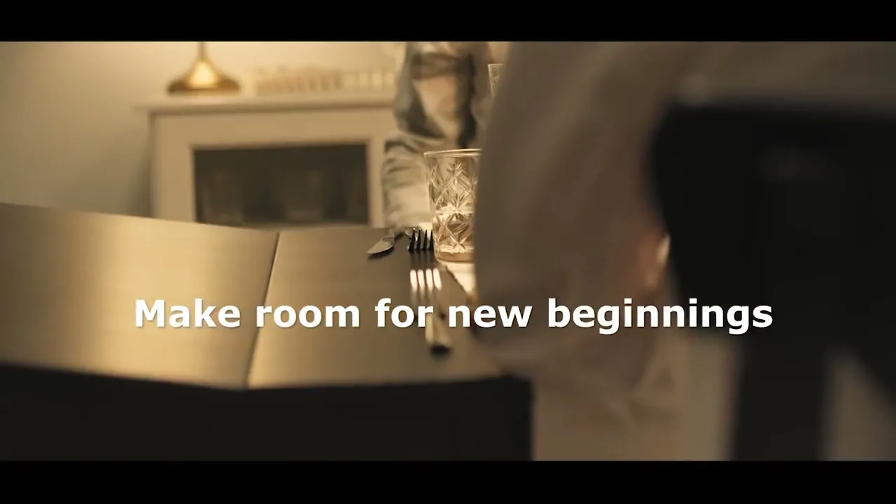 A person in a white shirt sets a dining table with cutlery and a glass, preparing for a meal in a warm, inviting room.