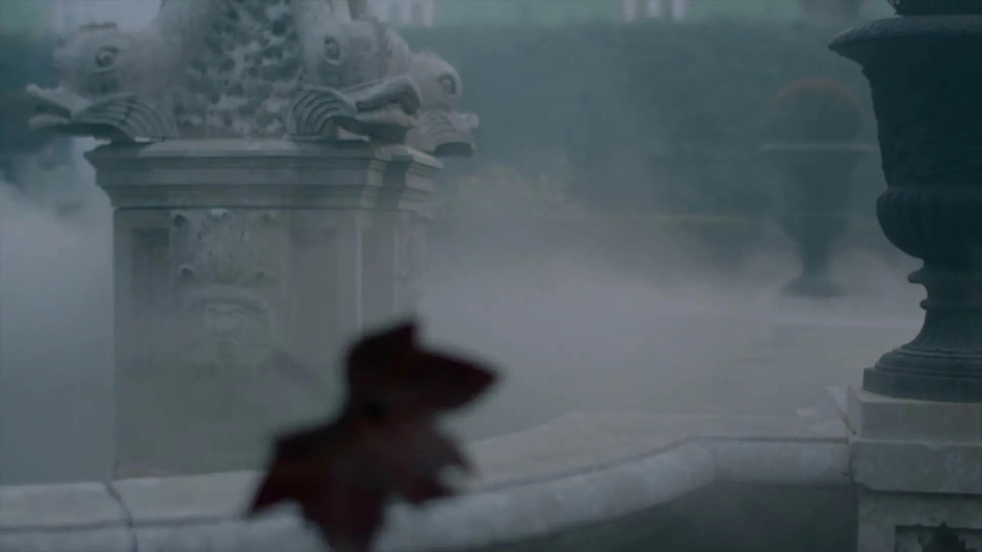 A stone fountain with carved faces and shells is partially obscured by mist, with a blurred red leaf in the foreground.