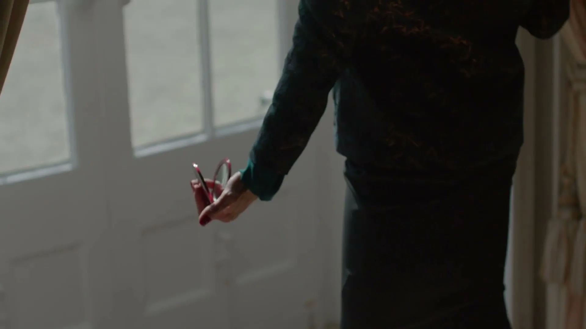 A woman in a dark green dress holds a small compact mirror in her hand, standing near a white door with glass panes.