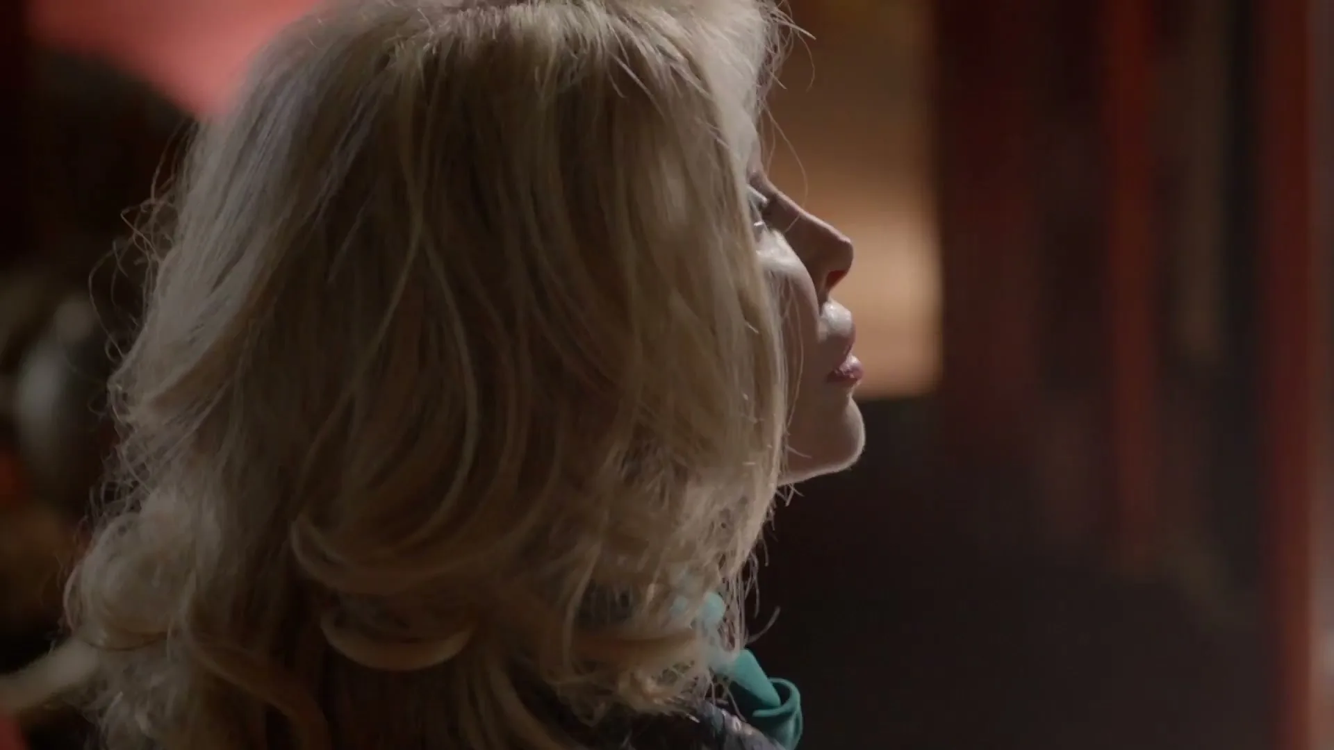 A blonde woman with voluminous hair looks up and to the right, her face partially illuminated by a soft light.