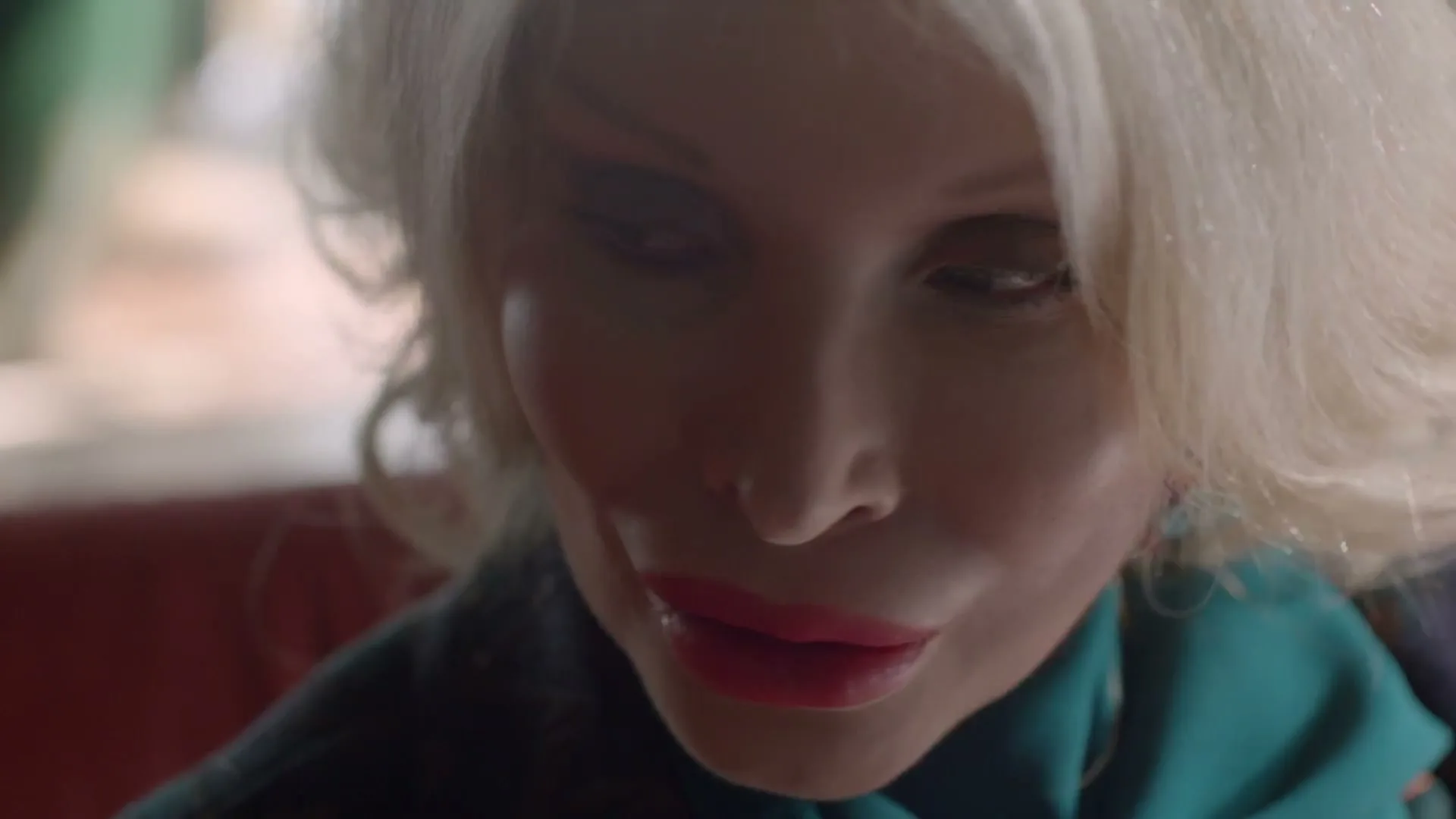 A blonde woman with full lips and heavy makeup looks down and to the side, speaking softly in an interior setting.