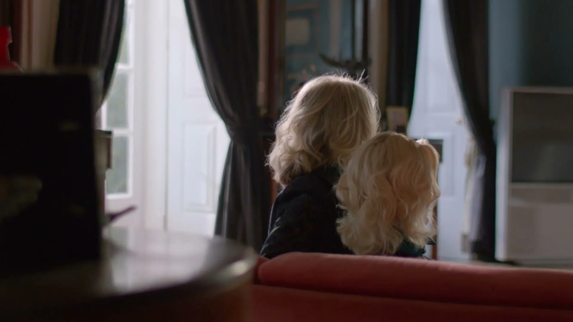 A woman with blonde hair holds a blonde doll, both facing away from the camera, looking out a window in a dimly lit room.