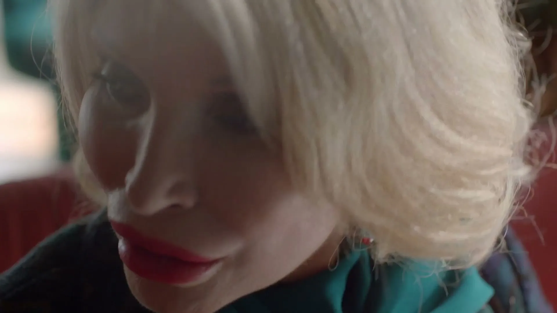 A woman with blonde hair and red lipstick looks down, wearing a teal scarf and a colorful earring, in an interior setting.