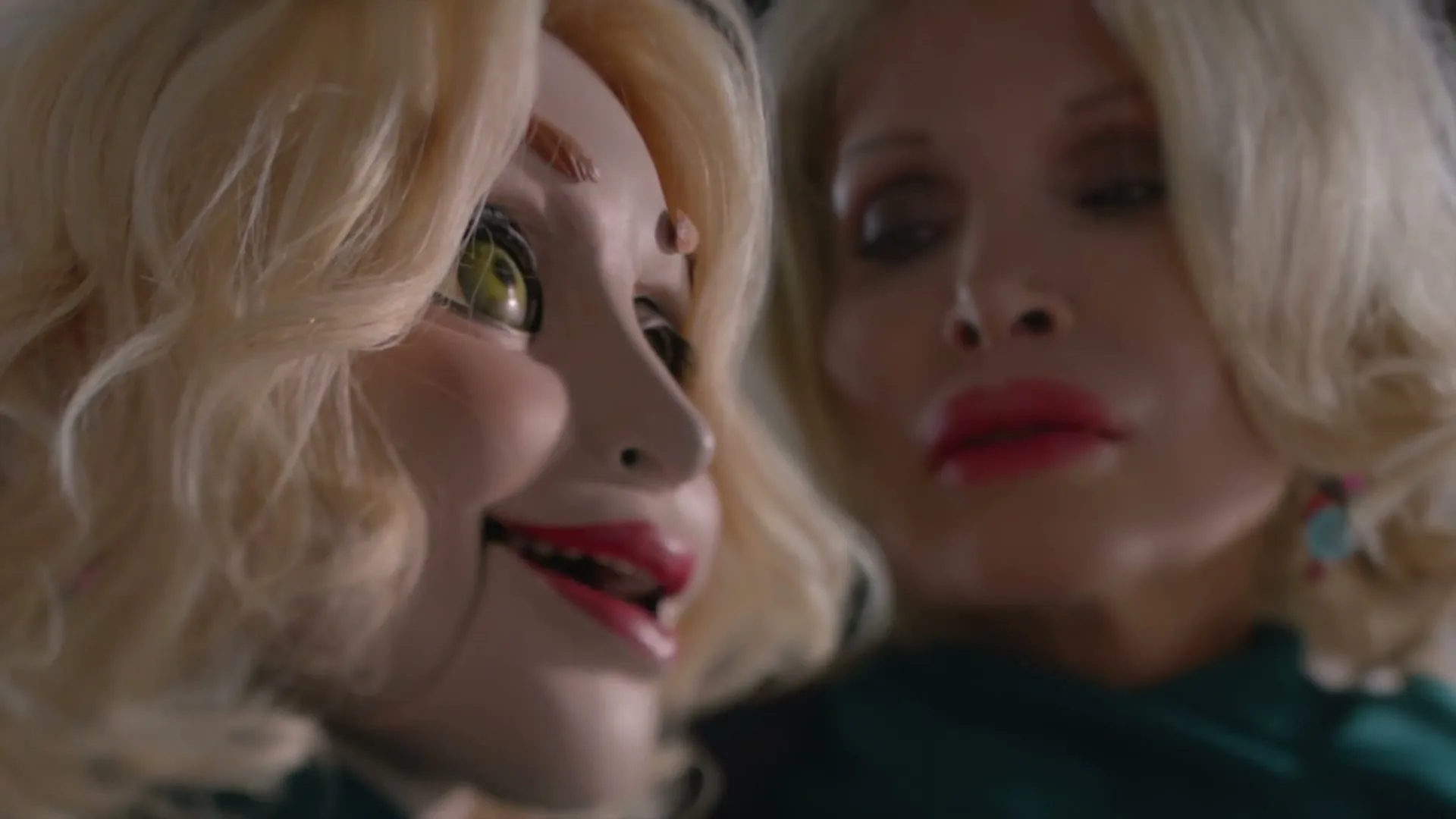 A blonde woman with exaggerated features and a doll-like face looks to the right, while another blonde woman with similar features is out of focus in the background.