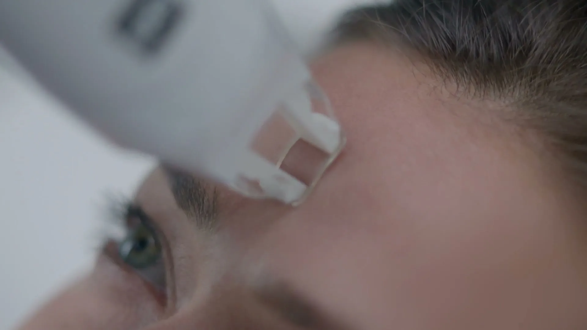 A close-up shot shows a woman's forehead as a white beauty device glides across her skin, performing a cosmetic treatment.