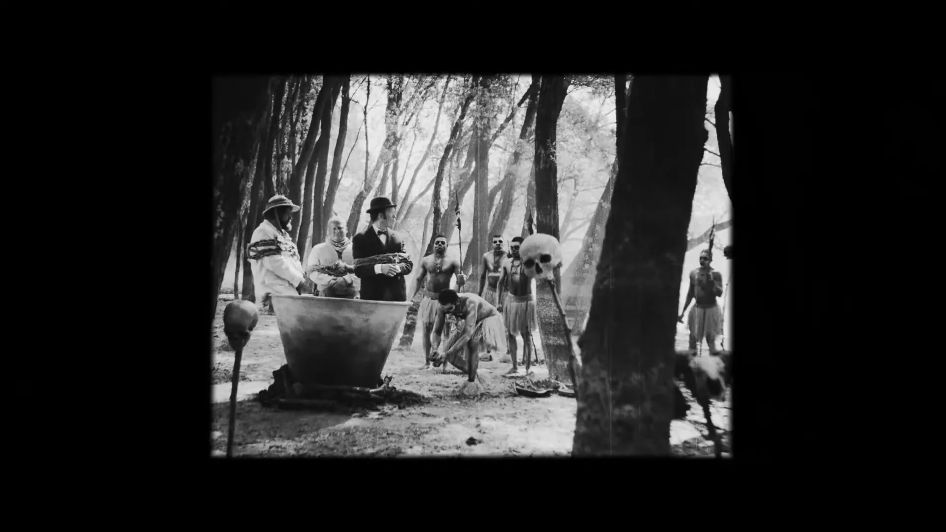 A man in a suit and bowler hat stands among a group of tribal people in a forest clearing, observing a large pot.