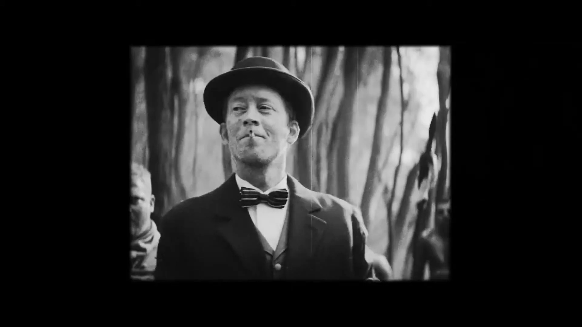 A man in a bowler hat and suit, with a cigarette in his mouth, stands tied up in a tub of water, surrounded by tribesmen in a jungle.
