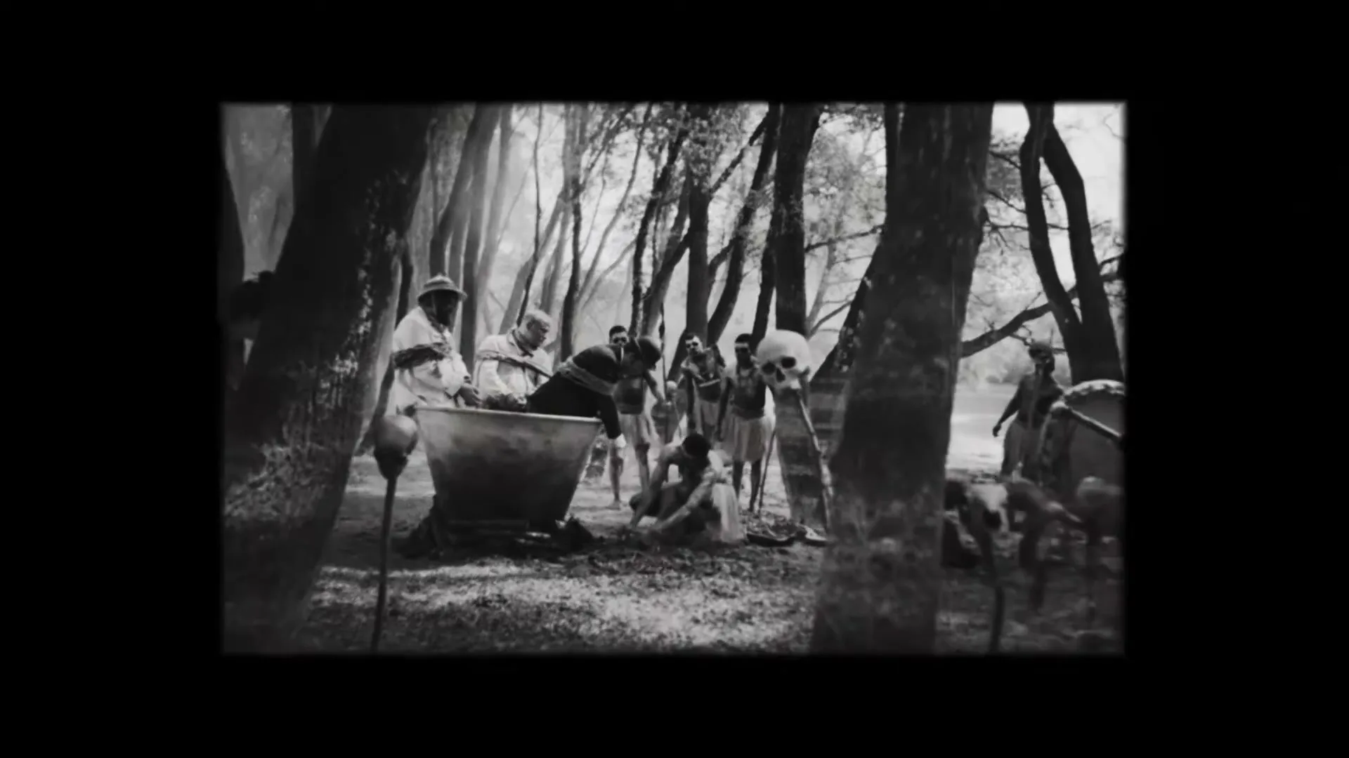 Two men, tied with ropes, are surrounded by indigenous people in a jungle clearing, as one man is lowered into a large pot.