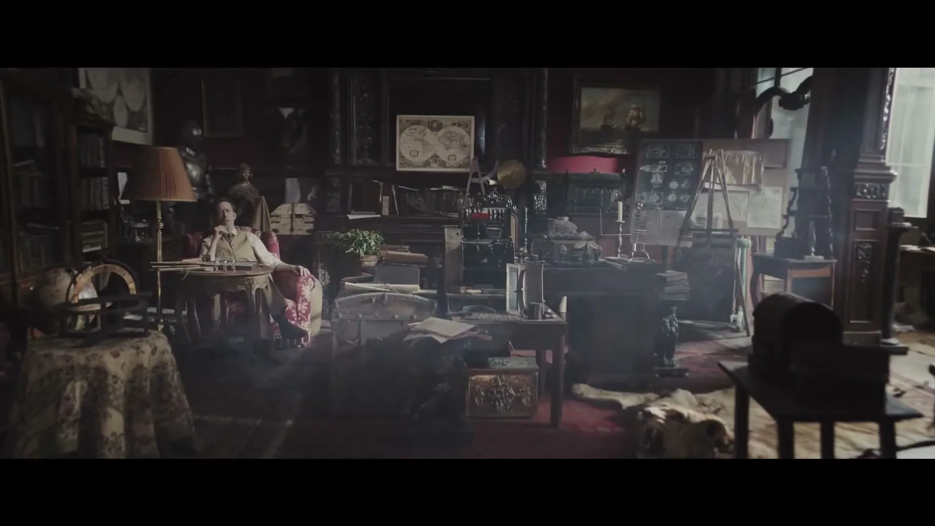 A man in a white shirt sits at a desk in a cluttered, dark, old-fashioned study, looking thoughtful.