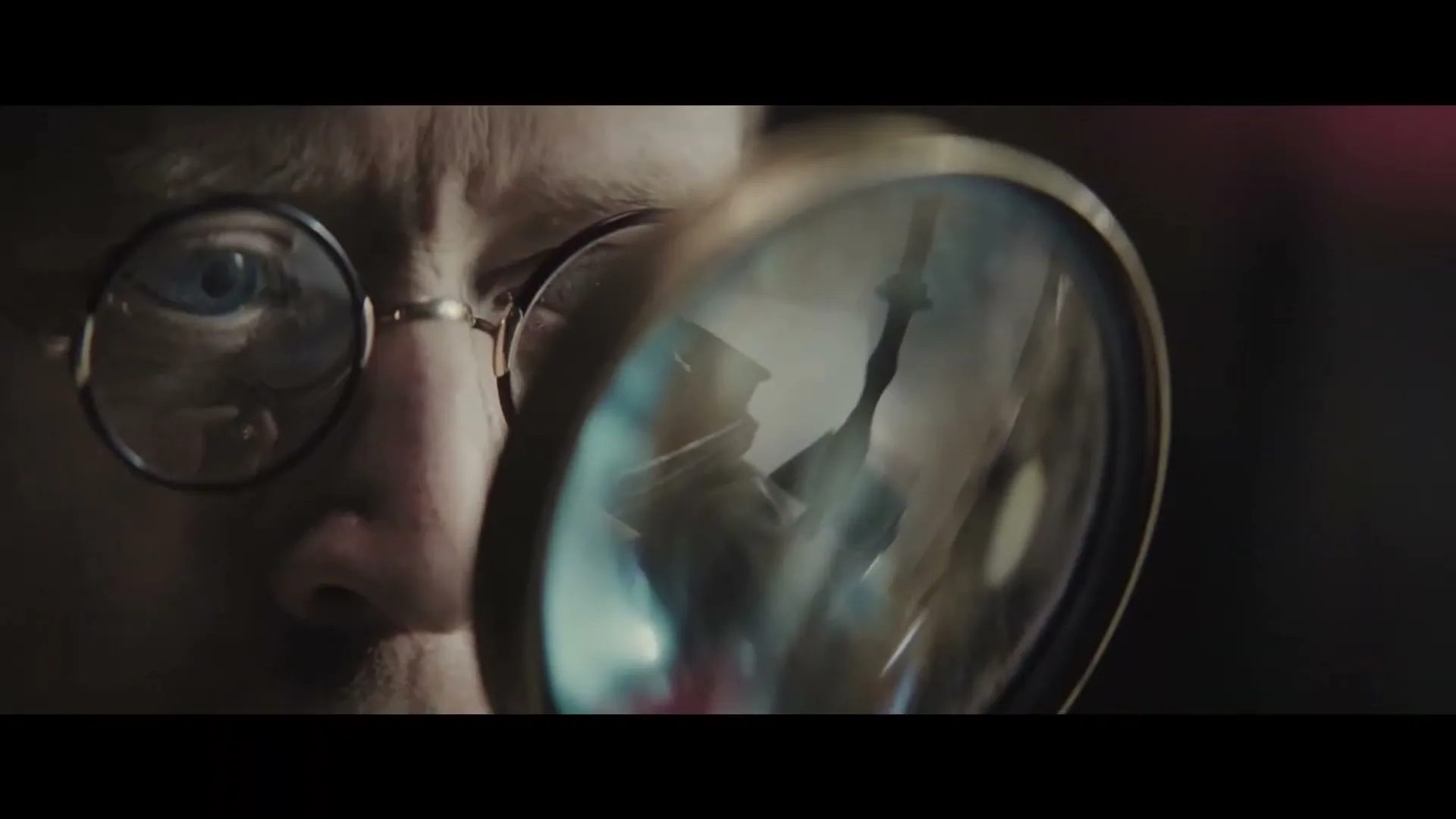 A man with glasses looks intently through a magnifying glass, reflecting a key, in a dimly lit interior.