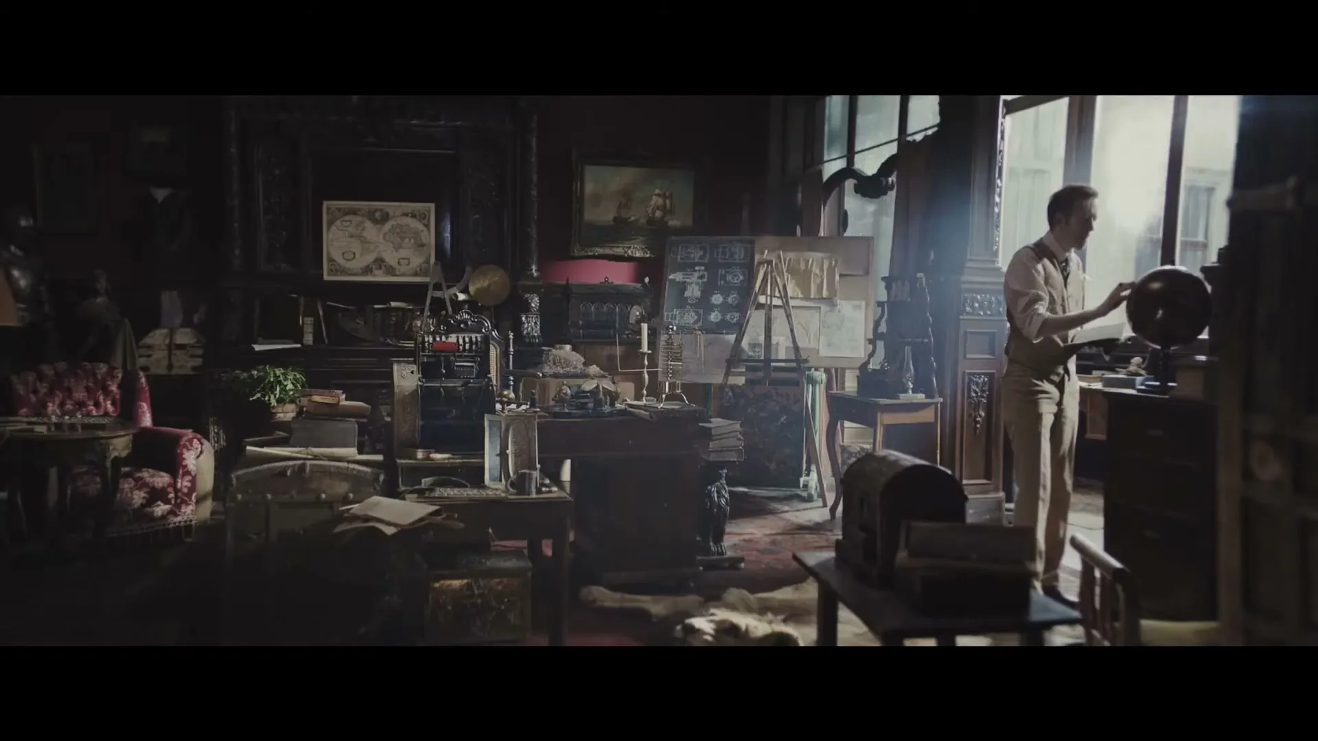 A man in his 40s, dressed in a vest and shirt, stands by a window in a cluttered study, looking at a globe.