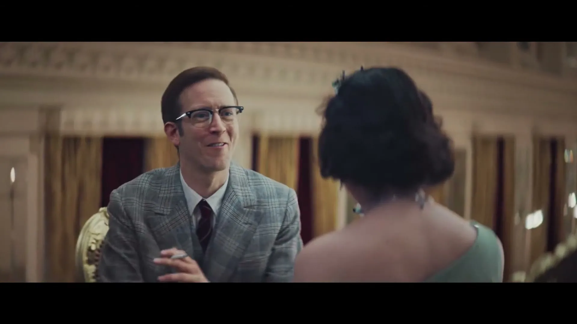 A man in glasses and a plaid suit smiles at a woman with her back to the camera in a grand, ornate room.