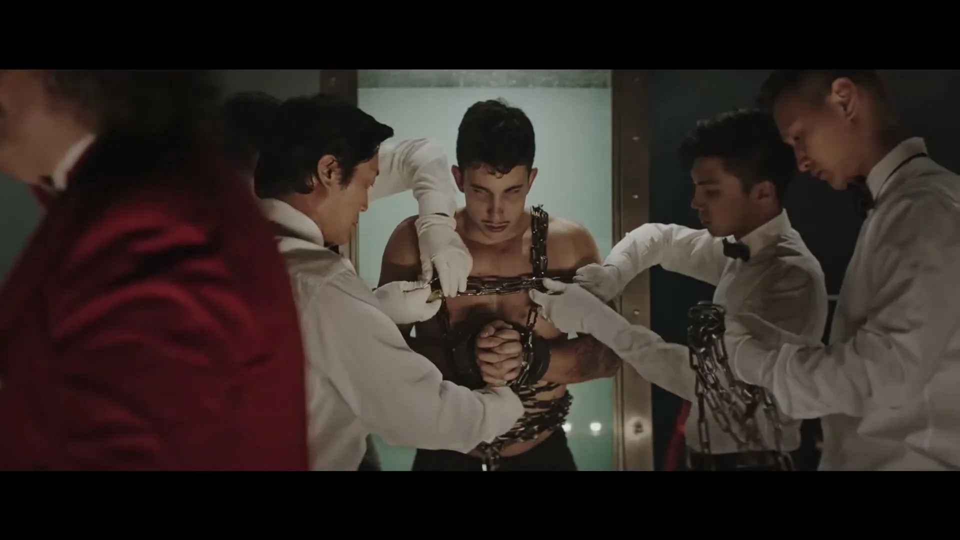 A muscular man is chained up by four men in white shirts and gloves, while another man in a red jacket watches in a dimly lit room.
