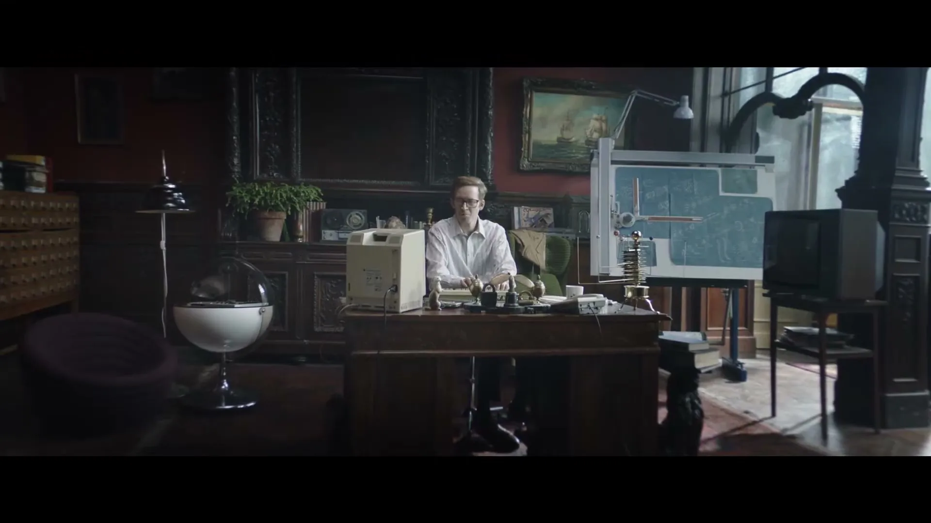 A man in his 30s sits at a large wooden desk in a dimly lit, ornate office, looking directly at the camera.