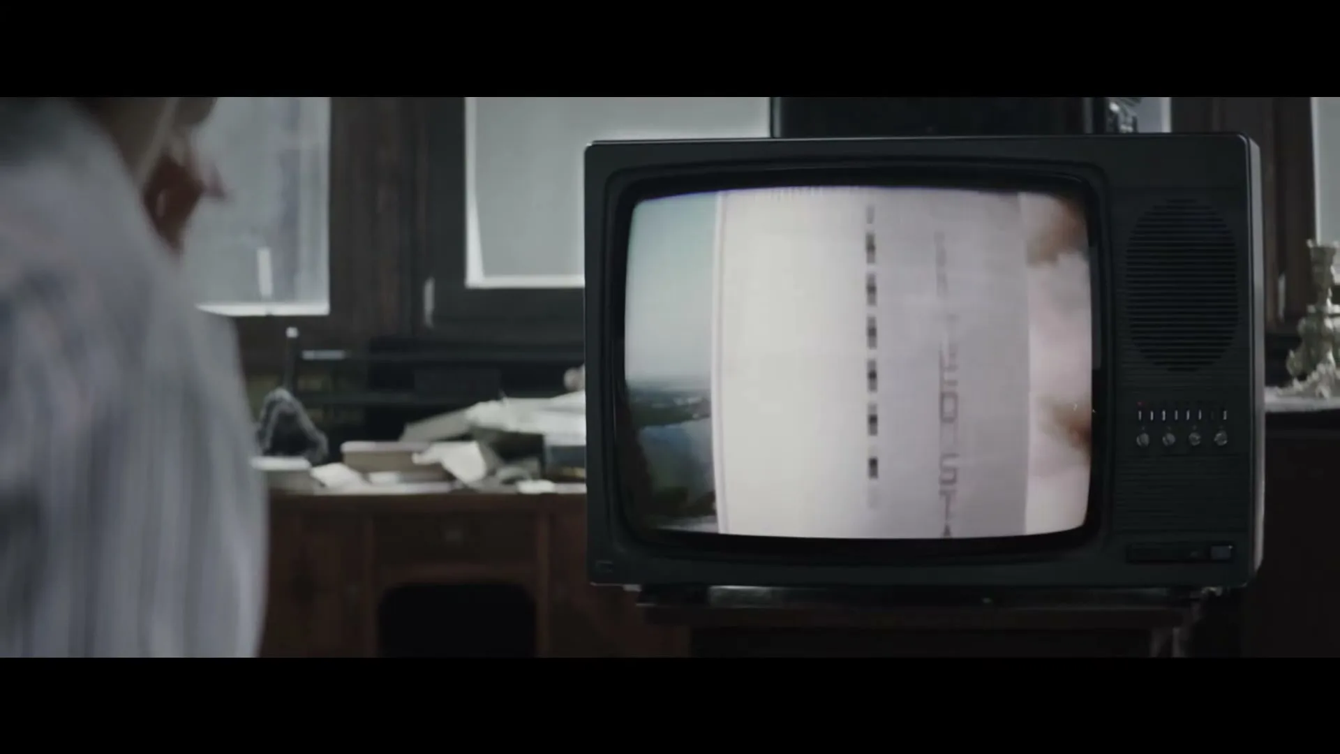 A man in a white shirt watches an old television set, which displays rapidly changing archival footage of historical events, in a dimly lit room.