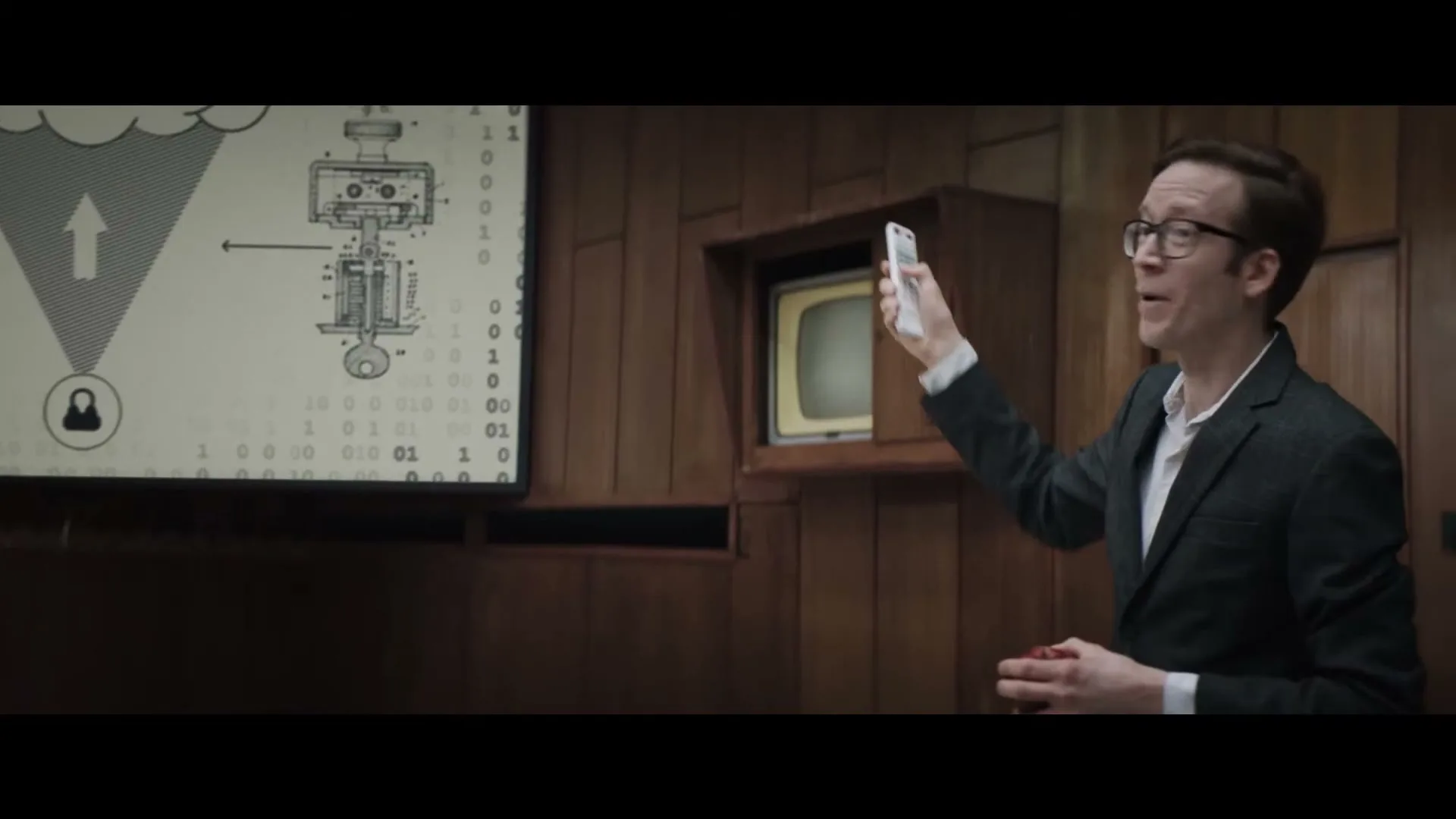 A man in his 30s, wearing glasses and a suit, points a remote control at a screen displaying a technical diagram in a wood-paneled room.