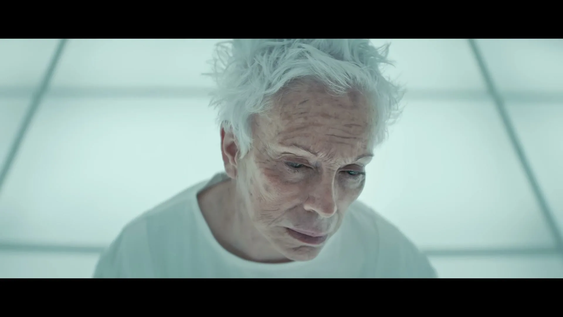 A woman with white hair and a white gown slowly opens her eyes, looking up in a sterile, white room.